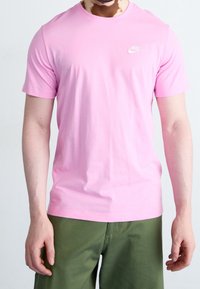 Vaaleanpunainen puuvillapaita lyhyillä hihoilla, pyöreä kaula-aukko ja pieni valkoinen Nike-logo rinnassa. Yhdistetty oliivinvihreisiin housuihin.