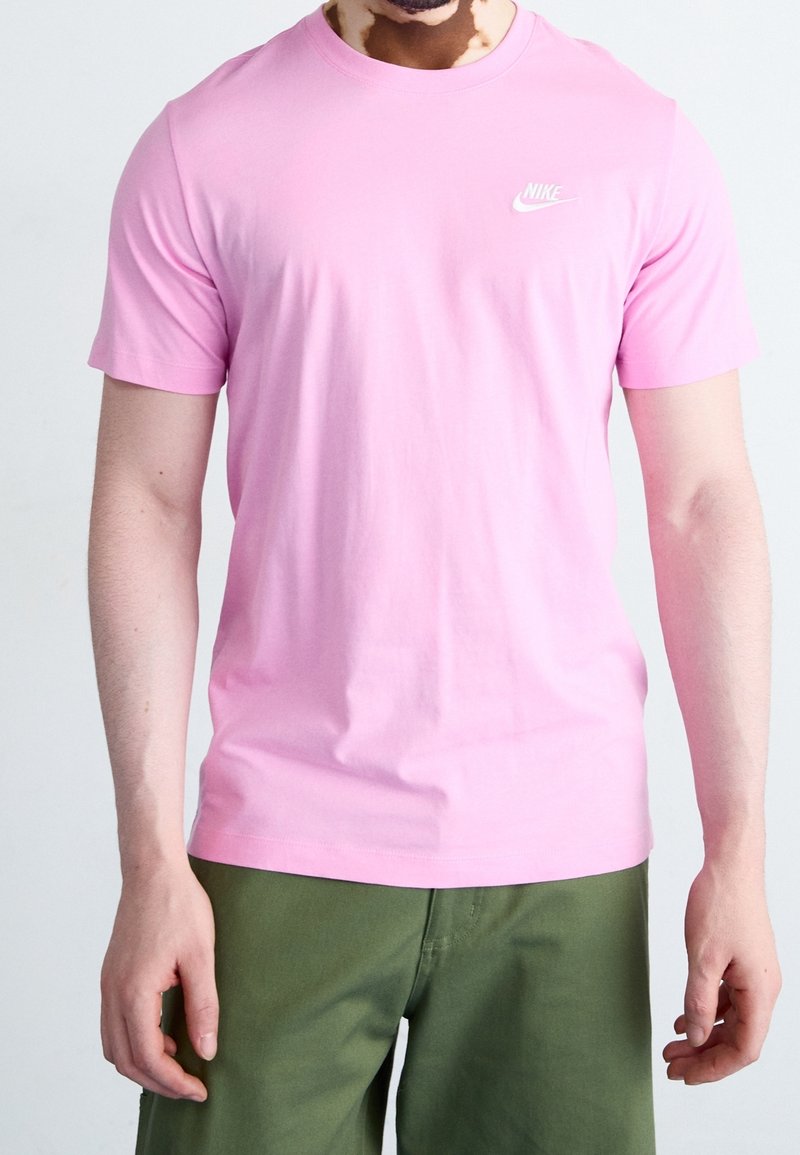 Vaaleanpunainen puuvillapaita lyhyillä hihoilla, pyöreä kaula-aukko ja pieni valkoinen Nike-logo rinnassa. Yhdistetty oliivinvihreisiin housuihin.