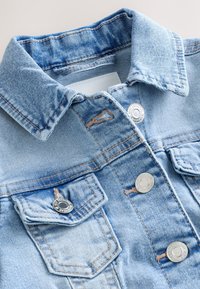 Giacca di denim azzurro chiaro con colletto piegato, due tasche frontali con patta e bottoni a pressione argento su sfondo bianco.