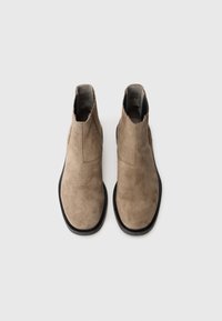 Un par de botas Chelsea de ante beige con suelas negras, vistas desde arriba sobre un fondo blanco liso.
