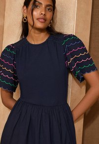 Abito blu navy con maniche a palloncino, dotato di ricami colorati a scollop. Tessuto liscio, vita arricciata e silhouette rilassata.