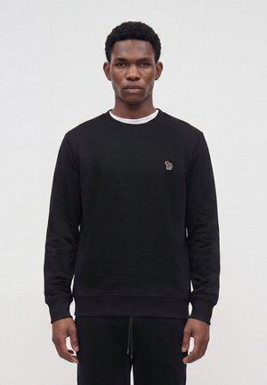 Homme portant un sweat-shirt noir à col rond avec un petit zèbre brodé sur la poitrine gauche, debout devant un fond gris uni.