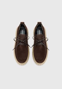 Des chaussures en daim marron avec un bout arrondi, des lacets plats et une semelle intérieure texturée marron, présentant une semelle extérieure en caoutchouc léger avec un motif de traction.