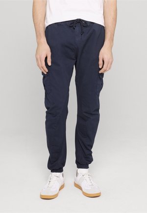 Man draagt marineblauwe cargo joggerbroek met elastische boorden, witte sneakers en een wit shirt, staand tegen een effen lichte achtergrond.