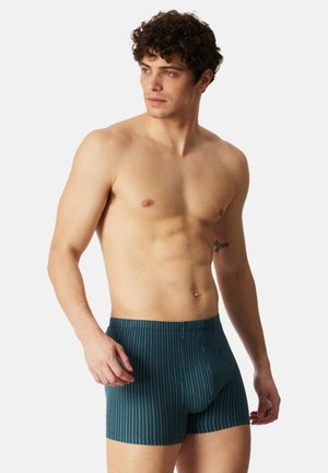 Ung mand med krøllet hår iført turkisblå boksershorts med striber, stående mod en ensfarvet lys baggrund og kigger til siden.