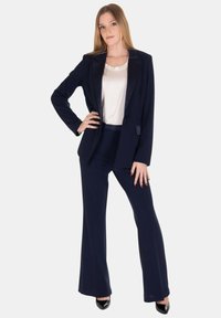 Completo blu navy composto da un blazer su misura e pantaloni a zampa, abbinato a una blusa crema. Il blazer presenta rever in satin e due tasche.