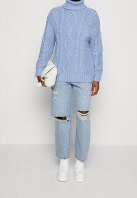 Maglione a collo alto lavorato a maglia blu chiaro, vestibilità rilassata, abbinato a jeans blu chiaro strappati e scarpe da ginnastica bianche con suola spessa. Borsa a mano bianca in mano.