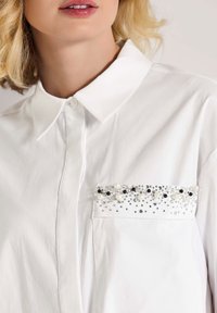 Weißes Hemd mit Button-Down-Kragen, ausgestattet mit einer Brusttasche, die mit schwarzen, weißen und silbernen Perlen und Perlen verziert ist.