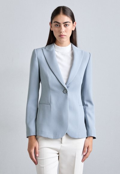 Lichtblauw op maat gemaakte blazer met een geknipte revers, enkele knoopsluiting en twee voorzakken, gecombineerd met een witte top met hoge hals.