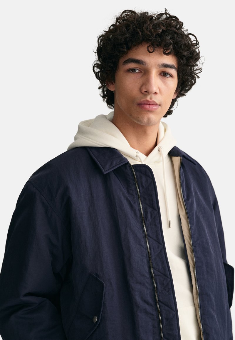 Navyblauer Nylonjacke mit einem Reißverschluss vorne, gestepptem Futter und seitlichen Taschen, die über einem weißen Kapuzenpullover getragen wird. Verfügt über einen Kragen und ein minimalistisches Design.