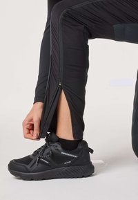 Person, der lynlåser benet på sorte atletikbukser over en sort vandtæt Swedemount-sneaker med synlige sorte sokker.