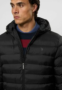 Schwarze Daunenjacke mit Reißverschluss, ge quilteter Textur und verstellbarem Kapuzen. Mit einem dezenten Logo auf der Brust und einem geschichteten Farbdesign darunter.