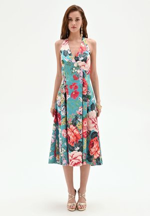 Donna che indossa un vestito estivo blu senza maniche con grandi stampe floreali multicolori, braccialetti dorati e sandali dorati con tacco e cinturini.