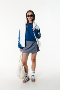 Maglione blu con stampe grafiche, gilet in pelliccia sintetica bianca, gonna a righe grigie, calze a righe bianche e blu, sneakers marroni e borsa in rete.