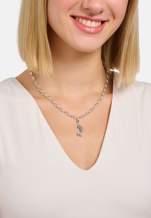 Lächelnde Frau mit blondem Haar, die eine silberne Halskette mit einem kleinen, fischförmigen Anhänger trägt und ein weißes V-Ausschnitt-Oberteil anhat.