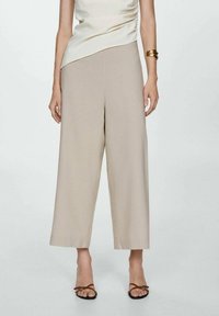 Žena v béžových širokých kalhotách do půli lýtek, krémovém tílku bez rukávů, hnědých sandálech na řemínky a zlatém náramku na pravé paži.