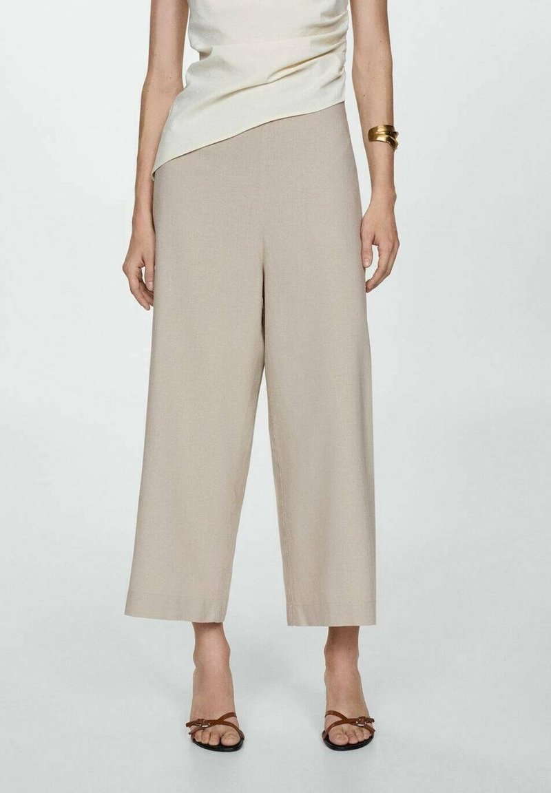 Žena v béžových širokých kalhotách do půli lýtek, krémovém tílku bez rukávů, hnědých sandálech na řemínky a zlatém náramku na pravé paži.