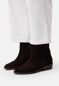 Stivaletti in suede marrone con punta affusolata e ritagli in stile western. Presentano una cerniera laterale e un tacco basso a tronchetto. Abbinati a pantaloni bianchi.