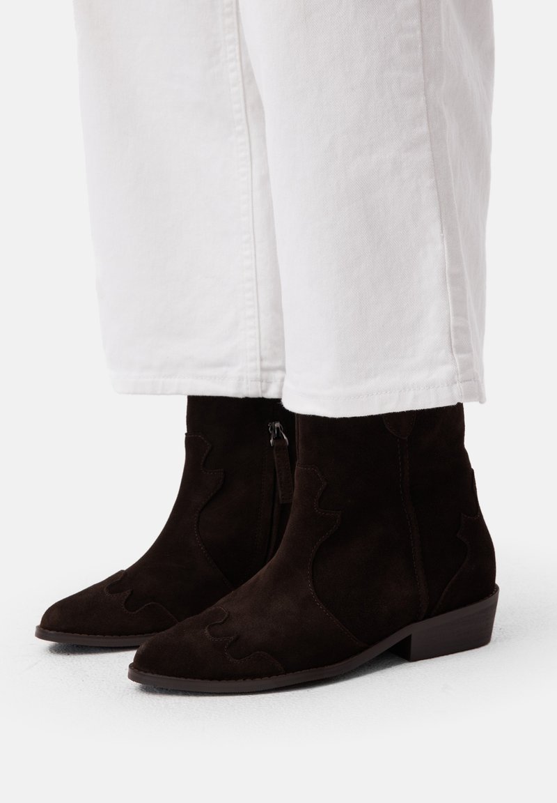 Braune Wildlederschnürstiefel mit spitzer Zehenform und ausgeschnittenem Western-Design. Mit einem seitlichen Reißverschluss und niedrigem, gestapeltem Absatz. Kombiniert mit weißen Hosen.
