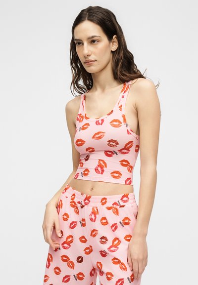 Γυναίκα που φοράει ροζ crop top και παντελόνι με κόκκινο και πορτοκαλί μοτίβο με αποτυπώματα χειλιών, όρθια με το ένα χέρι στην τσέπη μπροστά σε απλό φόντο.