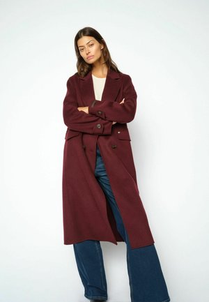 Kvinde stående med armene over kors iført en lang burgundy frakke, hvid top og vide blå jeans mod en ensfarvet hvid baggrund.
