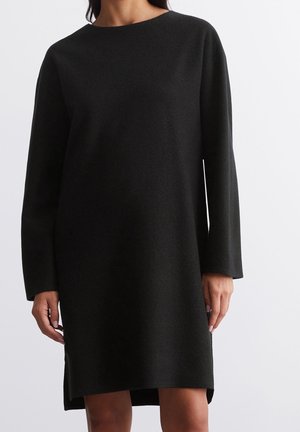 Vrouw die een zwarte wollen jurk draagt met lange mouwen en een lengte tot aan de knieën, met een ronde halslijn, tegen een effen achtergrond.