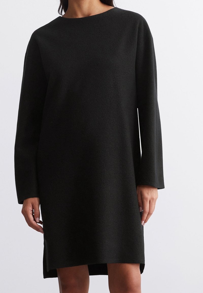 Femme portant une robe noire en laine à manches longues, longueur au genou, avec un col rond, sur un fond uni.