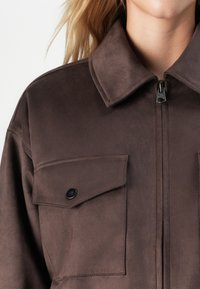 Jachetă din suede maro cu buzunar mare cu clapetă și nasture negru pe piept, închidere cu fermoar, purtată de o persoană cu părul blond.