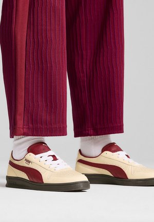 Sneaker bianca con dettagli rossi e suola grigio scuro, abbinata a pantaloni bordeaux a righe sottili, con calzini bianchi a coste.