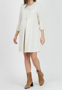 Nainen, joka on pukeutunut polven mittaiseen, kermanväriseen corduroymekkoon, jossa on kolme neljännestä hihoista, ja tan-värisiin mokkanahkasaappaisiin, joissa on paksut korot.