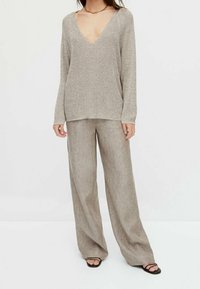 Beige gebreide trui met een diepe V-hals en lange mouwen, gecombineerd met lichtbruine wijde linnen broek. Platte zwarte sandalen maken de look compleet.