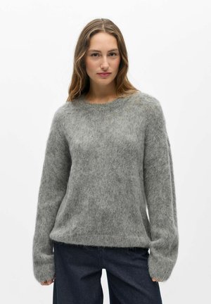 Jeune femme aux longs cheveux portant un pull doux gris pelucheux et un pantalon foncé, debout devant un fond blanc uni.