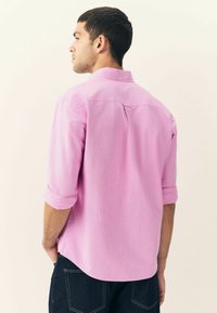 Camicia rosa con bottoni e maniche arrotolate, caratterizzata da una texture morbida e una leggera lucentezza. La parte posteriore include una piega centrale per un tocco di dettaglio in più.