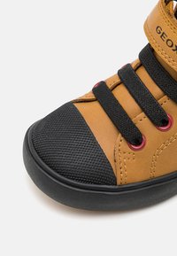 Zapatilla marrón con punta texturizada negra, cordones negros pasados por ojales rojos y suela de goma negra sobre fondo blanco.