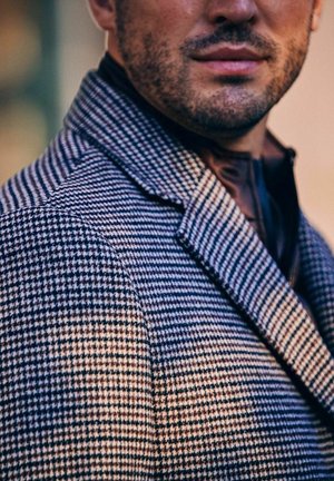 Baarddragende man met een jas met een houndstooth-patroon en een donker kledingstuk eronder, toont zijn ondergezicht en bovenlichaam in zacht licht.