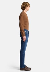 Brązowa koszula z długim rękawem zestawiona z niebieskimi dżinsami. Strój charakteryzuje się dopasowanym krojem i gładką fakturą, dopełniony brązowymi mokasynami.