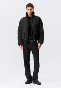 Schwarze Steppjacke mit seitlichen Taschen, hohem Kragen und elastischen Bündchen, darüber ein schwarzer Pullover, kombiniert mit dunklen Jeans und Schuhen.