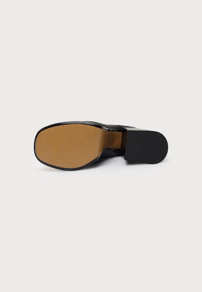 Chaussure habillée noire vue de dessous, avec une semelle beige et un talon noir solide, posée à plat sur un fond blanc.