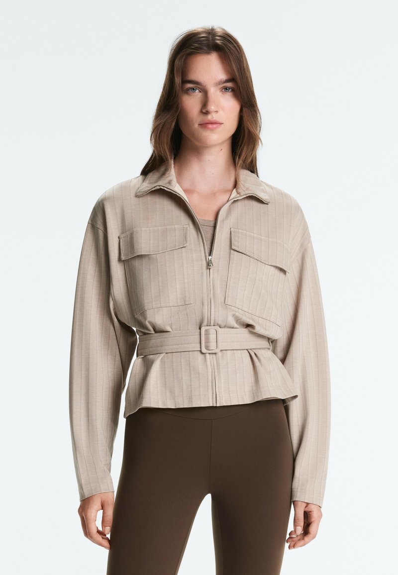 Femme portant une veste beige ceinturée avec des poches devant et un pantalon brun foncé ajusté, debout devant un fond clair uni.