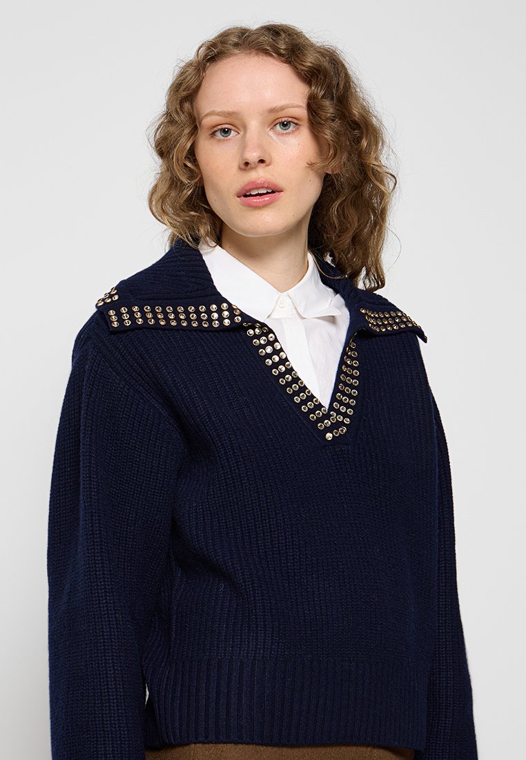 Pull en tricot bleu marine avec un décolleté en V profond, un col orné de clous dorés, des poignets et une taille côtelés. Tissu texturé.