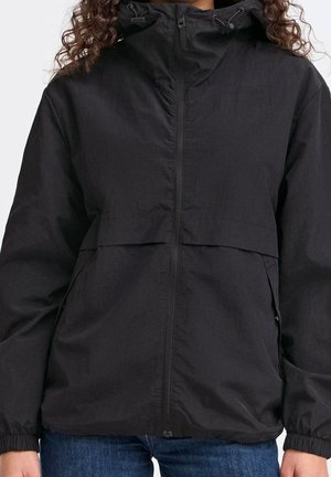 Personne portant une veste coupe-vent noire à capuche avec fermeture éclair à l'avant et poignets élastiques, associée à un jean bleu.