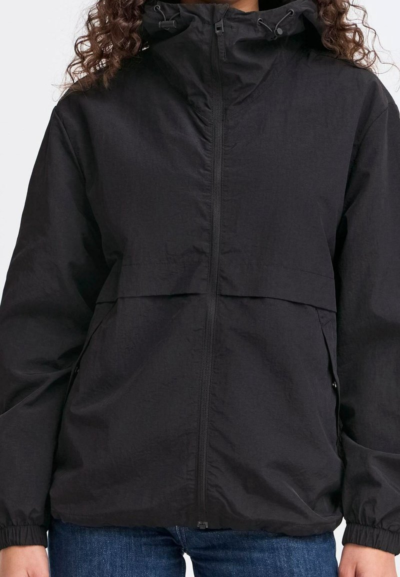 Personne portant une veste coupe-vent noire à capuche avec fermeture éclair à l'avant et poignets élastiques, associée à un jean bleu.