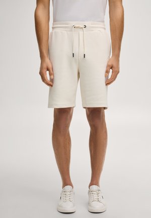 Mężczyzna ubrany w kremowe szorty ze sznurkiem, białe trampki i białą koszulkę bez rękawów, stojący na tle jednolitego tła.