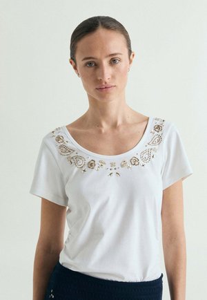 Joven mujer con cabello oscuro recogido en un moño bajo, que lleva una camiseta blanca de manga corta con bordados florales dorados y beige alrededor del escote redondo.