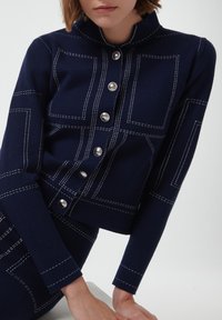 Chaqueta de punto en color azul marino con cuello alto, botones plateados y costuras blancas en un patrón geométrico. Diseño ajustado con mangas largas.