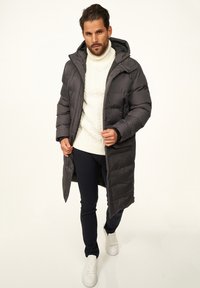 Langer Steppmantel in Dunkelgrau mit hohem Kragen und Kapuze. Getragen über einem cremefarbenen Kabelstrickpullover und marineblauen Hosen, kombiniert mit weißen Turnschuhen.