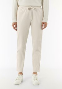 Helle beige, dehnbare Jogginghosen mit einem Kordelzugverschluss, Seitentaschen und schmal zulaufenden Beinen. Getragen mit weißen Turnschuhen vor einem neutralen Hintergrund.