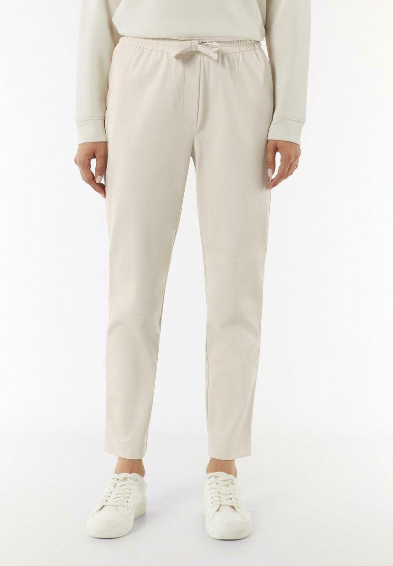 Helle beige, dehnbare Jogginghosen mit einem Kordelzugverschluss, Seitentaschen und schmal zulaufenden Beinen. Getragen mit weißen Turnschuhen vor einem neutralen Hintergrund.