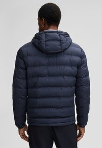 Marineblauwe pufferjack met capuchon, voorzien van gestikte secties, elastische manchetten en een glad, glanzend oppervlak. Achteraanzicht toont een minimalistisch ontwerp.