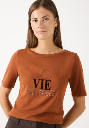Jonge vrouw met lang bruin haar die een roestkleurig shirt draagt met de tekst "La Vie Est Belle" in zwarte en strassletters.